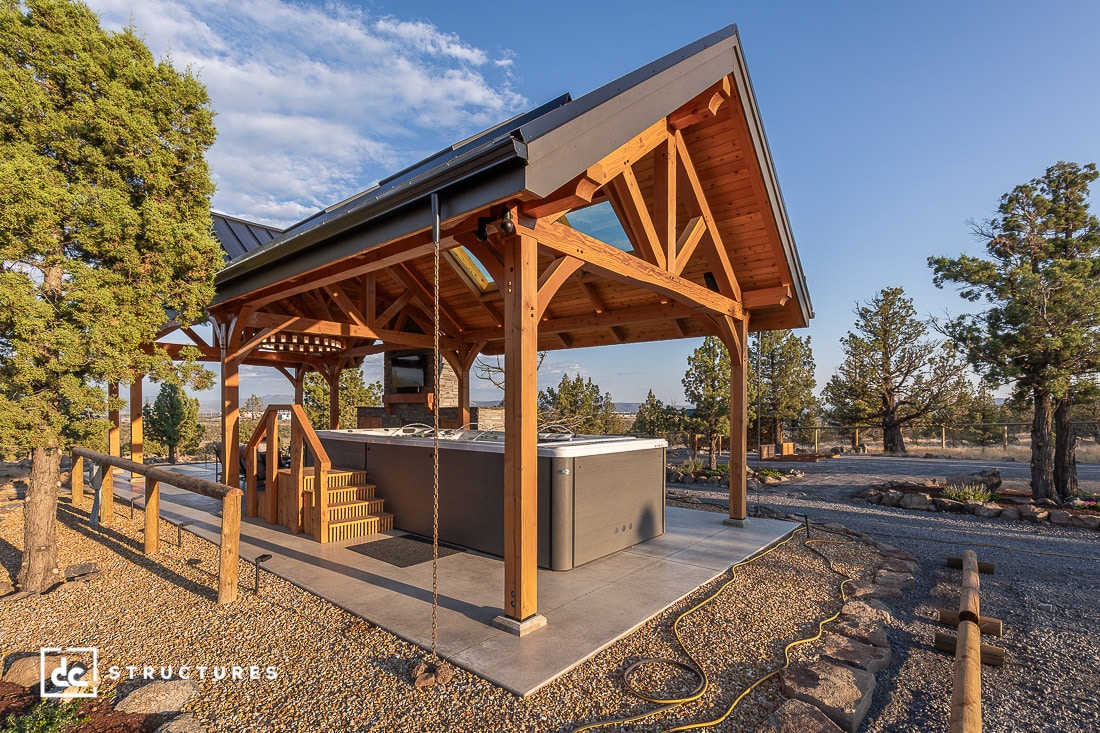 A wooden open-air pavilion with a peaked roof shelters a large hot tub and steps, set in a rustic outdoor area under clear skies.