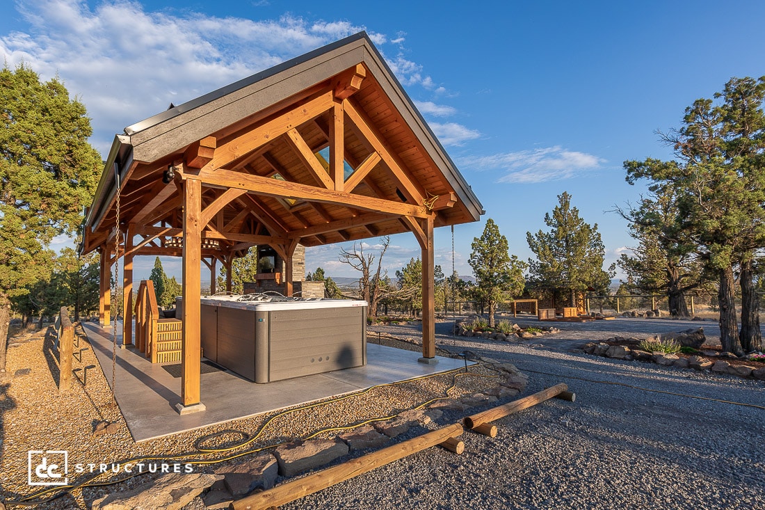 A wooden open-air pavilion shelters a hot tub on a concrete pad, surrounded by trees and gravel paths under a clear blue sky.