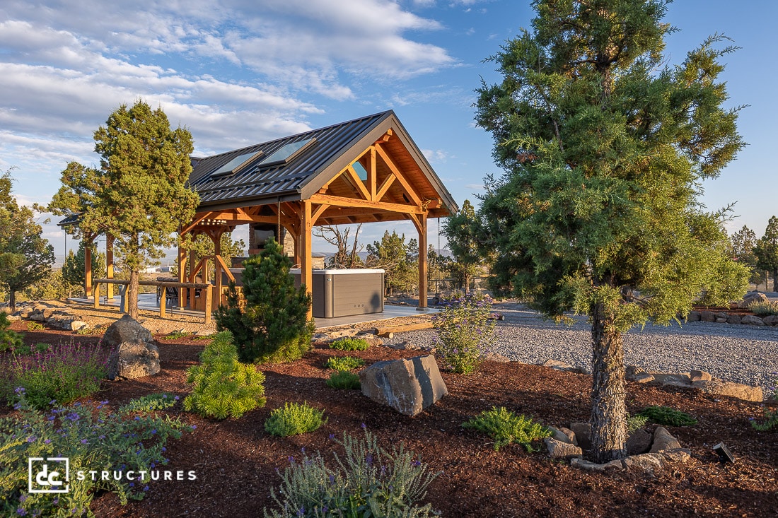 A wooden pavilion with a metal roof sits beside a hot tub, surrounded by landscaped gardens, trees, and gravel under a blue sky.