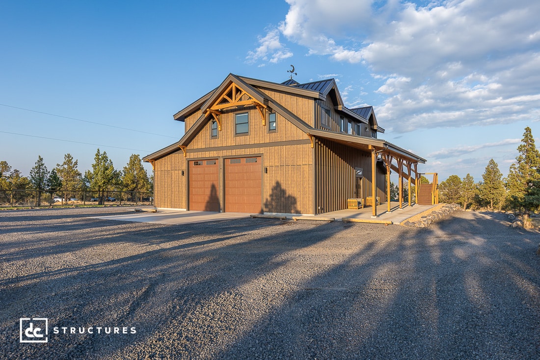 A modern barn-style building with wooden siding, double garage doors, and a covered porch sits on a gravel driveway under a blue sky.