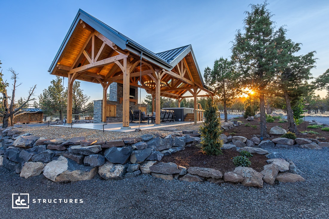 A wooden pavilion with a gabled roof on a stone foundation is surrounded by landscaping and trees at sunset, with outdoor furniture underneath.