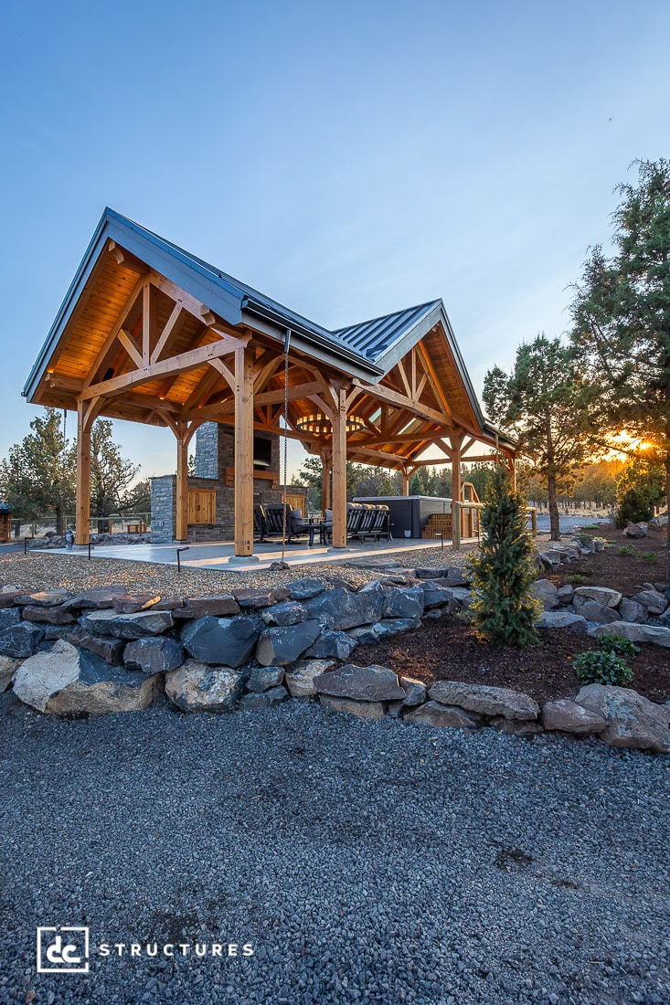 A wooden open-air pavilion with a metal roof stands on a stone-paved area, surrounded by rocks and small trees at sunset.