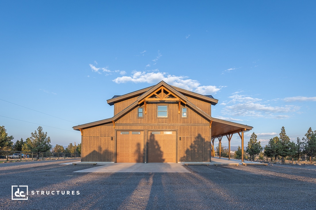 A large wooden barn-style building with two garage doors, a covered side porch, and a triangular roof stands on a gravel lot with trees and blue sky in the background.