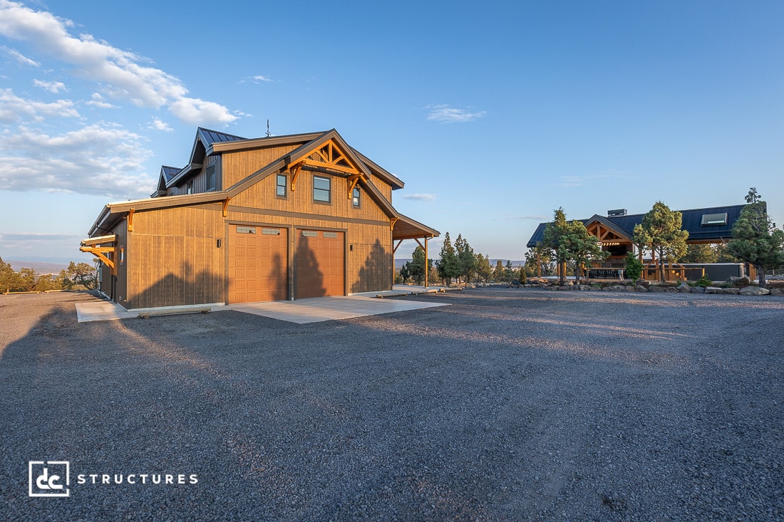 A large, modern barn-style building with wooden accents and two garage doors stands on a gravel lot under a clear sky.