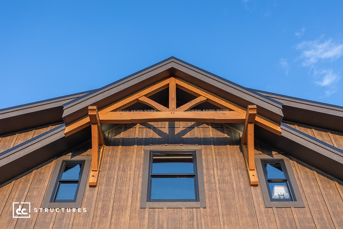 A close-up view of a modern wood-framed gable roof with exposed beams above three black-framed windows, under a clear blue sky.