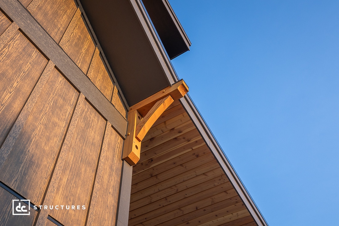Close-up of a wooden building's exterior corner, showcasing detailed wood grain, a decorative support bracket, and a sloped roof against a clear blue sky.