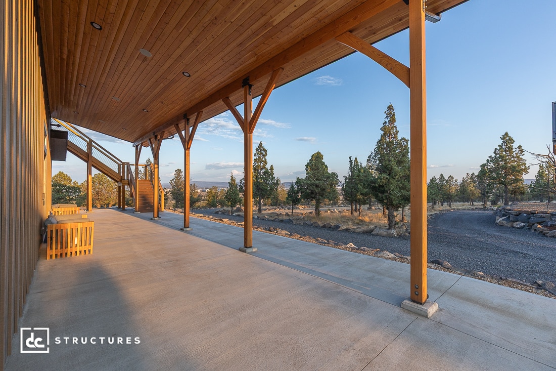 A wide wooden porch with a slanted roof overlooks a gravel driveway and scattered trees under a clear sky. Benches sit along the side, and sunlight casts shadows across the concrete floor.