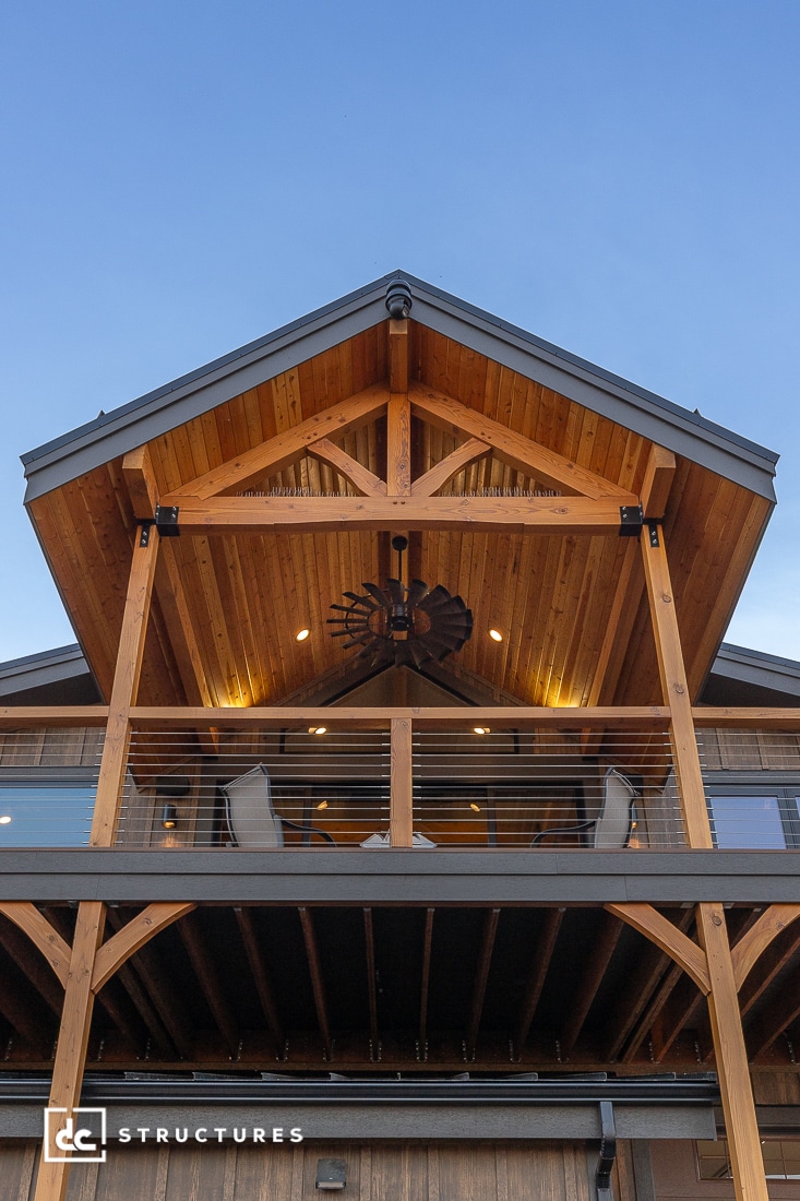 A two-story wooden house with a pitched roof, covered balcony, railings, patio furniture, and exposed beams. Warm lights above.