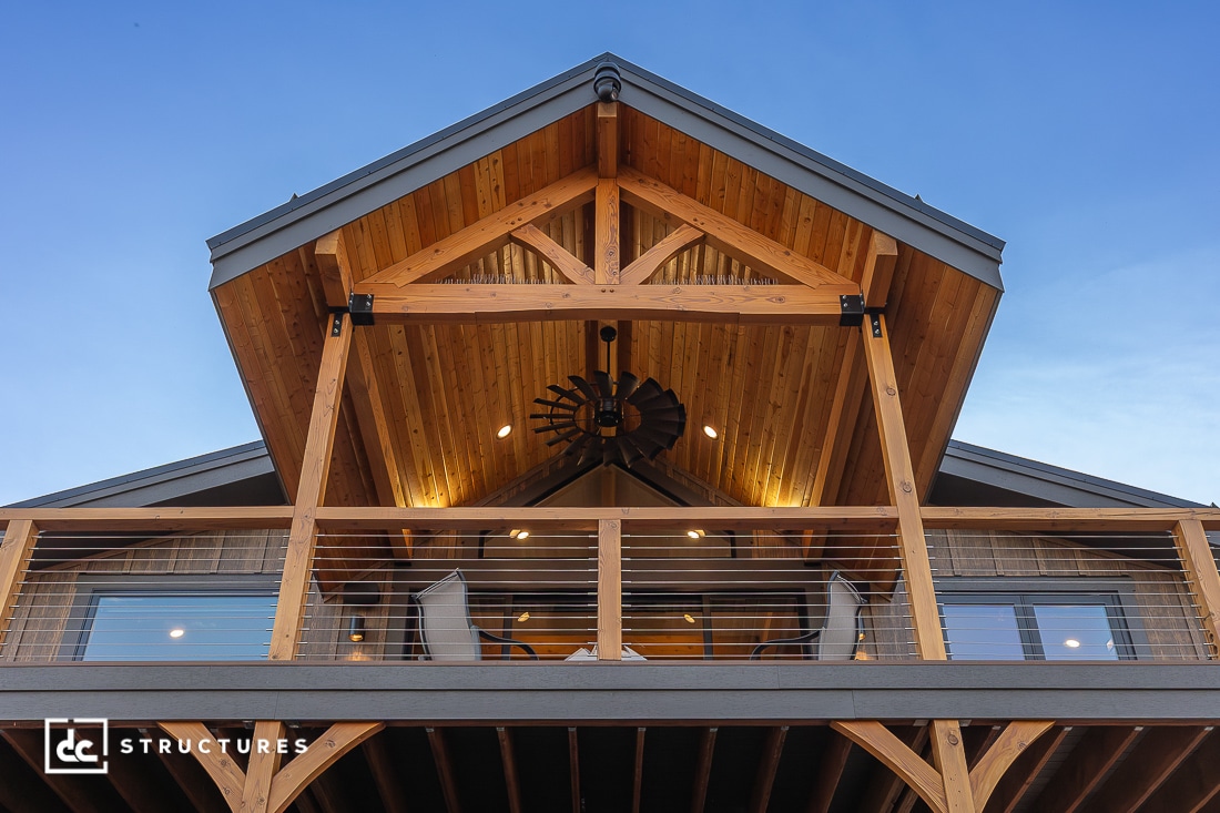 A modern wooden house with a steep gabled roof, exposed beams, and a covered balcony, photographed at dusk with a clear sky.