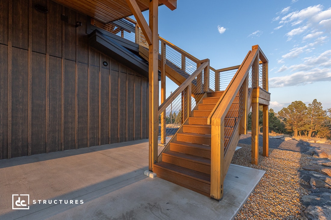 Exterior wooden staircase with metal cable railing attached to a modern building with vertical wood siding, overlooking a gravel landscape and trees at sunset.