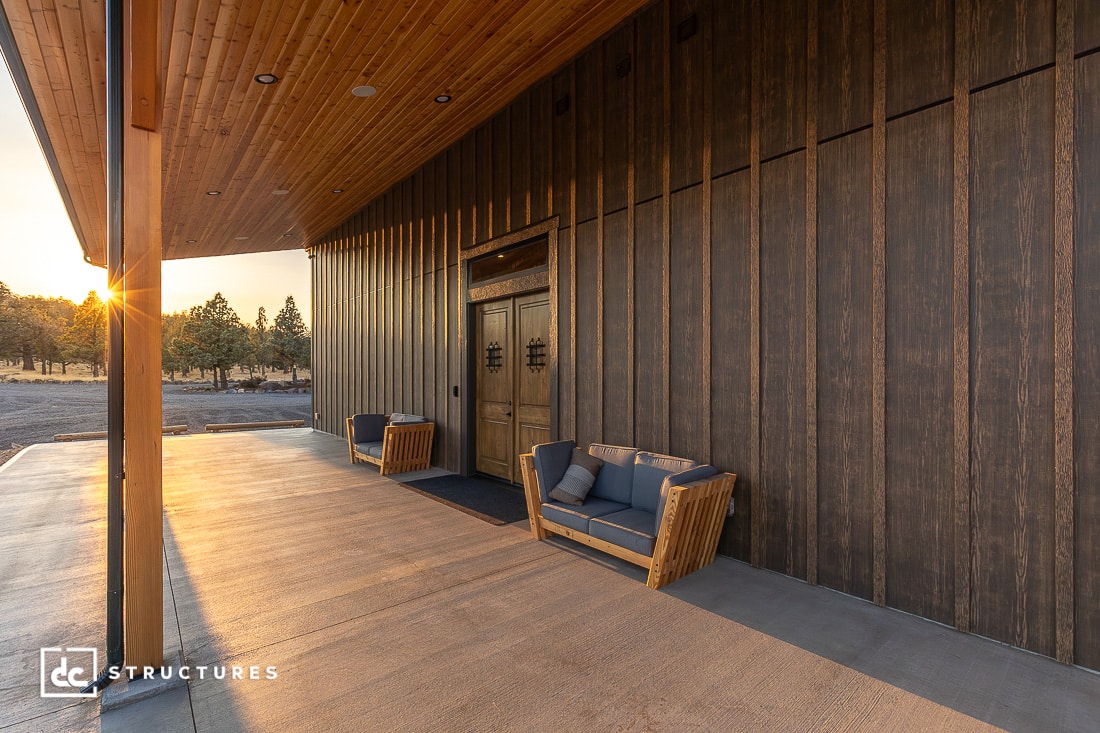 A modern wooden building with a covered patio, two wooden-framed sofas, and double front doors. Sunlight casts long shadows across the concrete, highlighting trees and open land in the background.