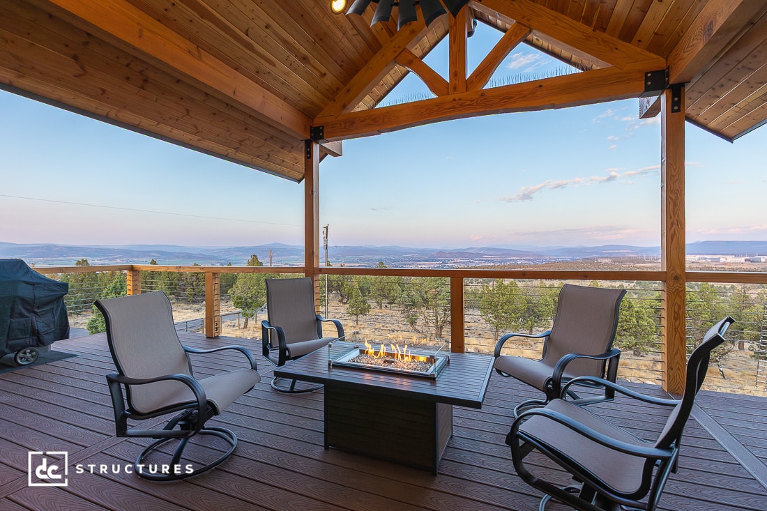 A covered wooden deck with four cushioned chairs around a square fire pit, overlooking trees and mountains at sunset.