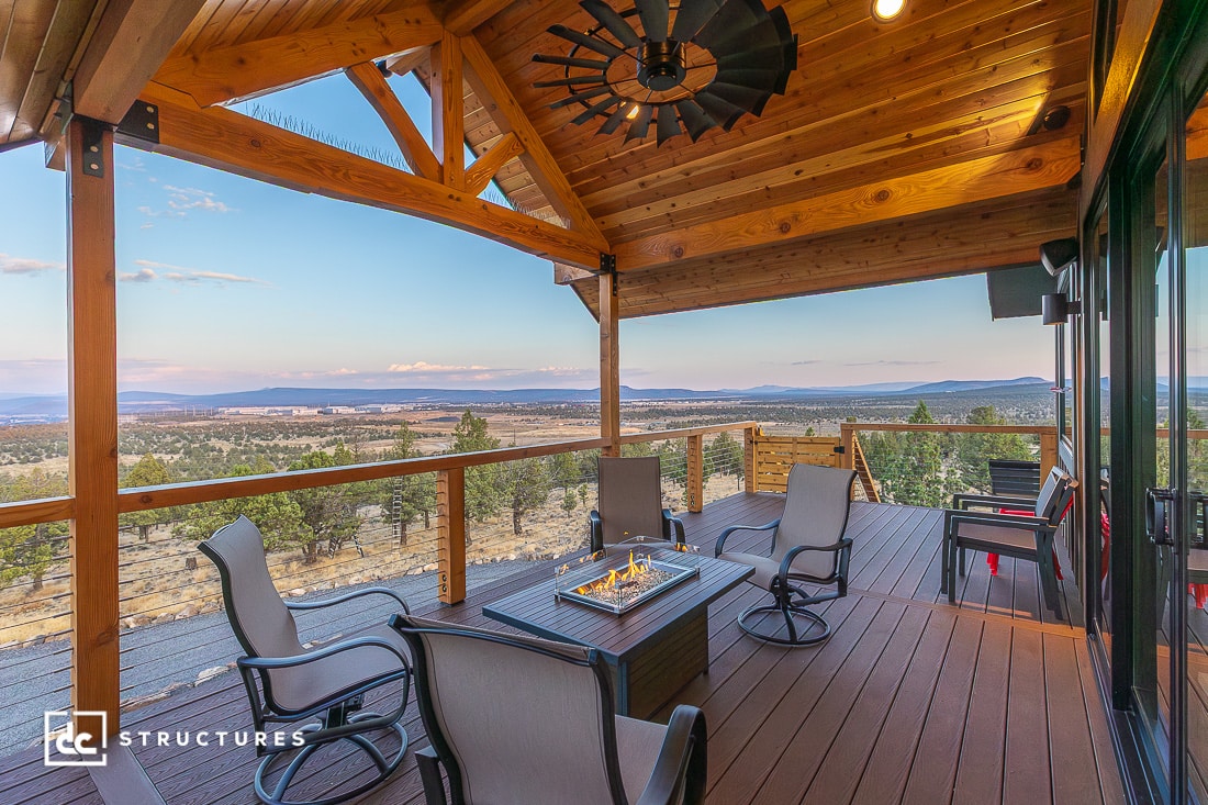 A wooden deck with a fire pit table, cushioned chairs, and railing overlooks trees and mountains under a partly cloudy sky.