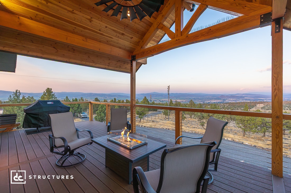 A covered wooden patio with four chairs around a fire pit, overlooking a valley and mountains at sunset; barbecue grill on the left.