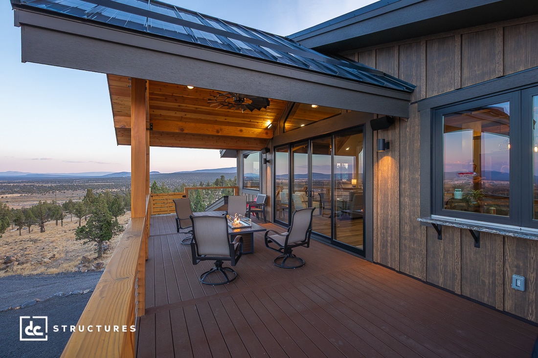 A spacious wooden deck with outdoor seating and a fire pit overlooks trees and hills at sunset. Glass doors connect to the house.