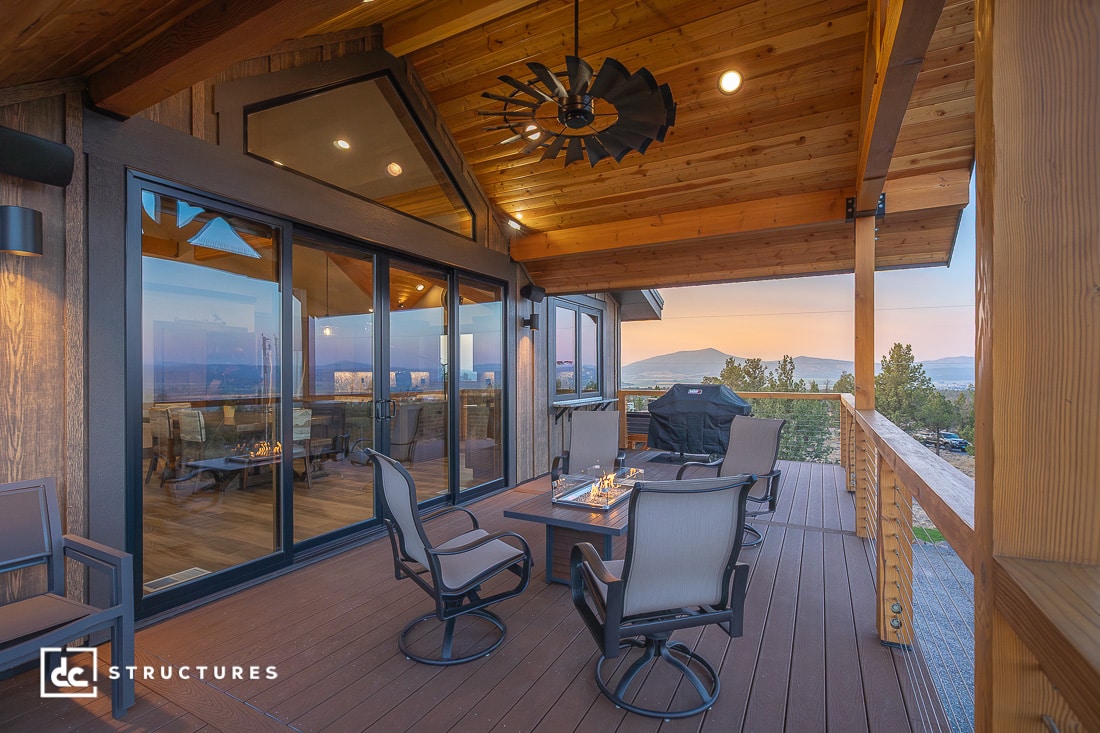 A covered wooden patio has modern outdoor seating, a fire pit table, a grill, and mountain views at sunset. Large glass doors lead inside, with a windmill-style ceiling fan above.