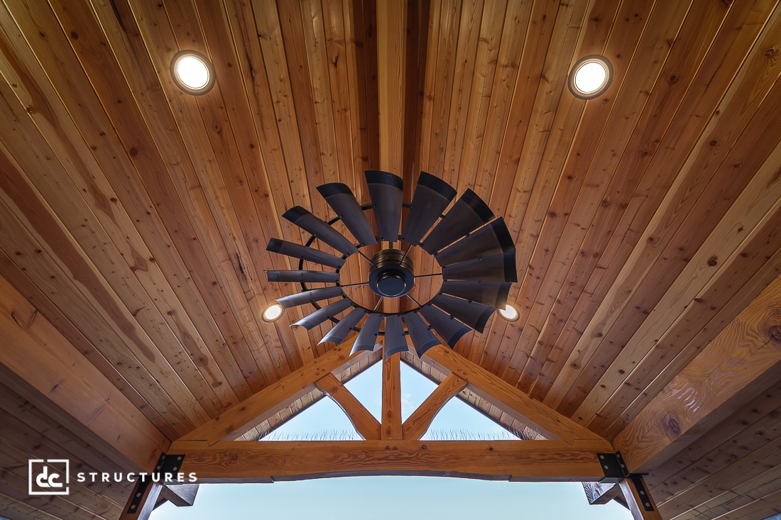 Ceiling view of a wooden structure with exposed beams and recessed lights, featuring a windmill-inspired ceiling fan in the center.