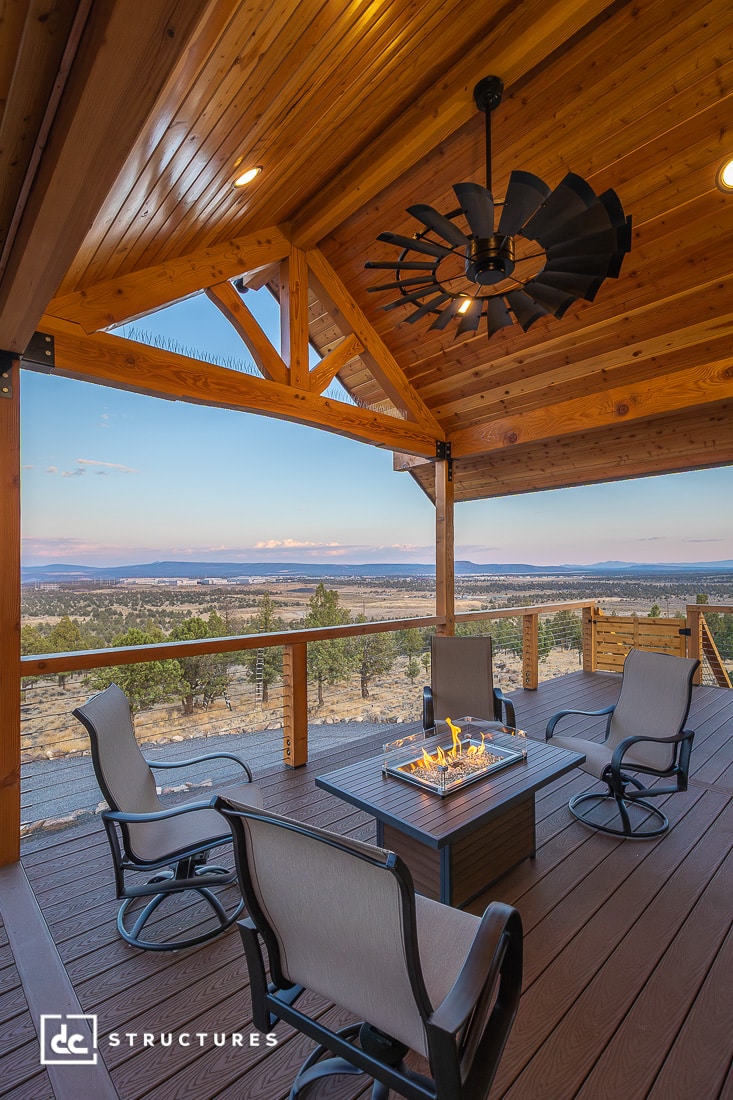 A covered wooden deck with four cushioned chairs around a fire pit table, ceiling fan above, and a view of fields, trees, and hills.