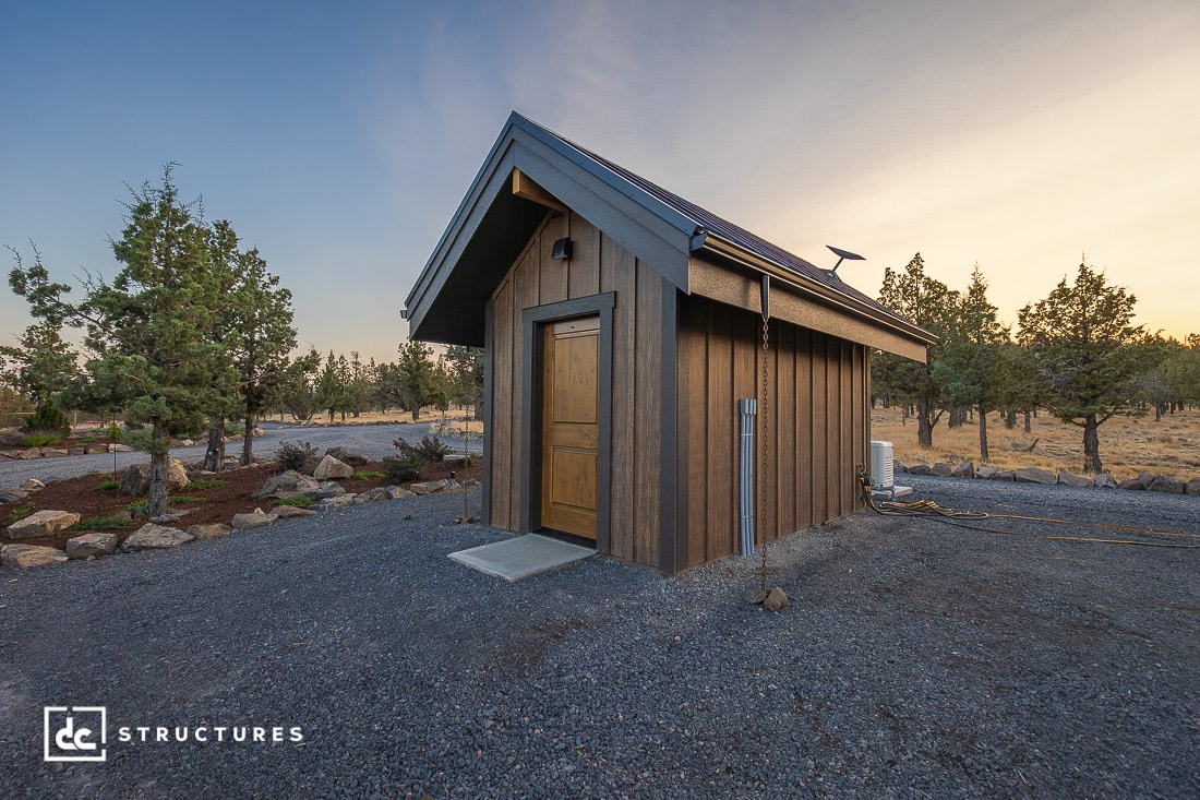 A small, modern wooden cabin with a pitched roof sits on gravel ground surrounded by trees. The sky is clear at sunset.
