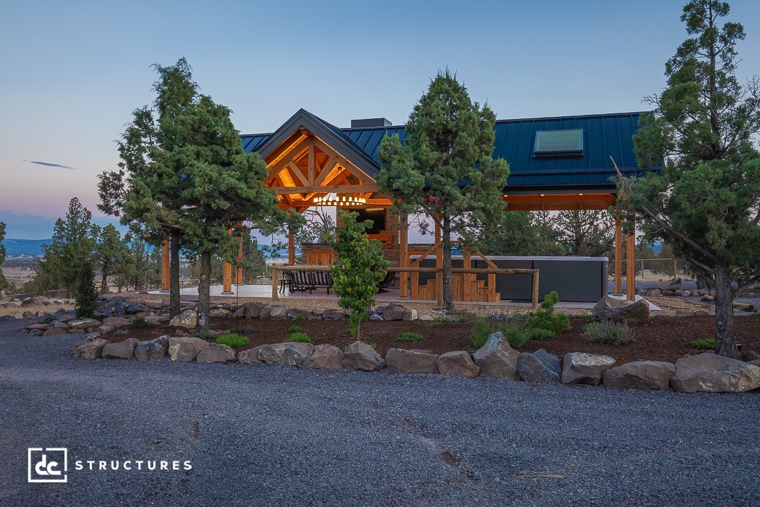 A modern wooden pavilion with exposed beams and a dark metal roof stands among trees, illuminated by warm lights at dusk.