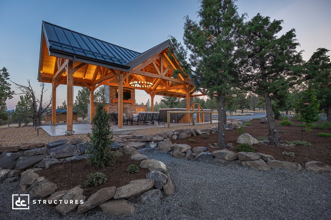 A wooden outdoor pavilion with a peaked metal roof stands among trees and rocks, warmly lit with a bar area and stools inside.