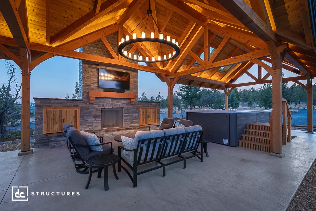 An outdoor covered patio with wooden beams, a circular chandelier, cozy seating by a stone fireplace, TV, and hot tub at dusk.