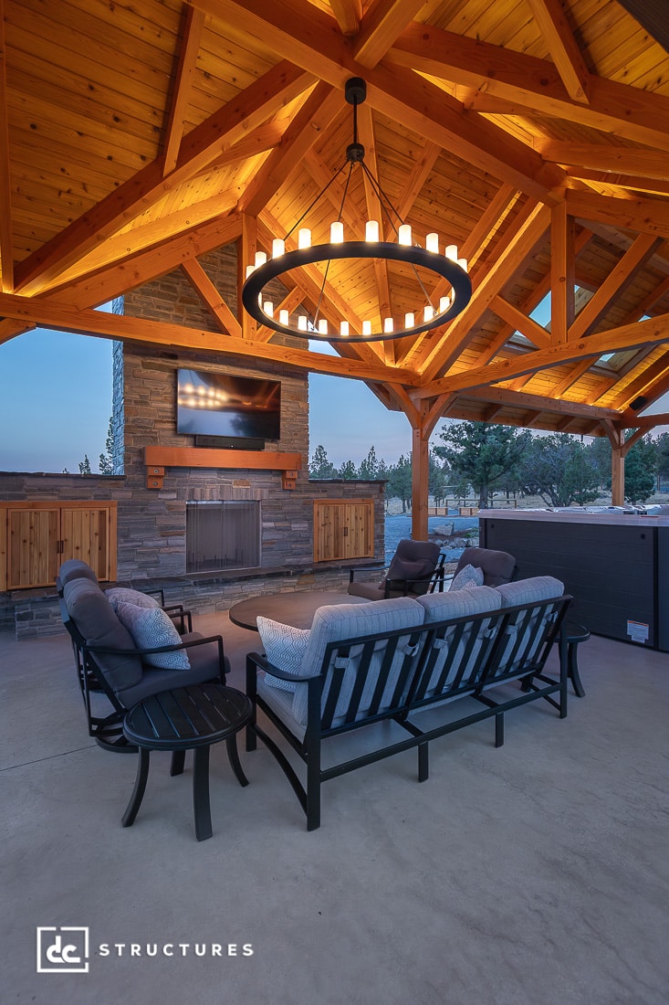 An outdoor seating area with cushioned chairs and a sofa under a pergola, modern chandelier, fireplace, and trees at dusk.