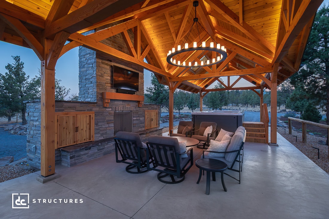 A covered outdoor patio with wooden beams, a stone fireplace, cozy seating, and a modern chandelier. Trees surround the space at dusk.