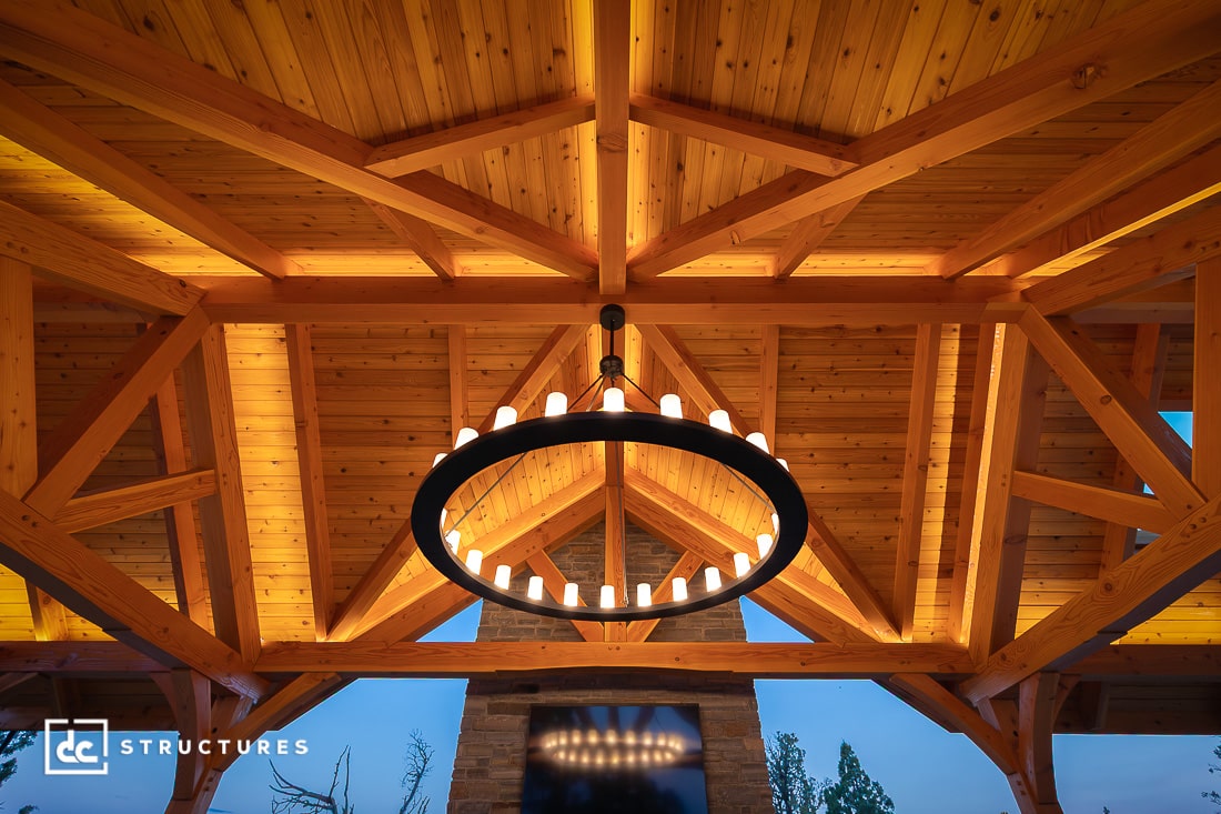 A circular chandelier with lit bulbs hangs from a vaulted wooden ceiling with exposed beams. A stone fireplace is below, and trees are visible outside.