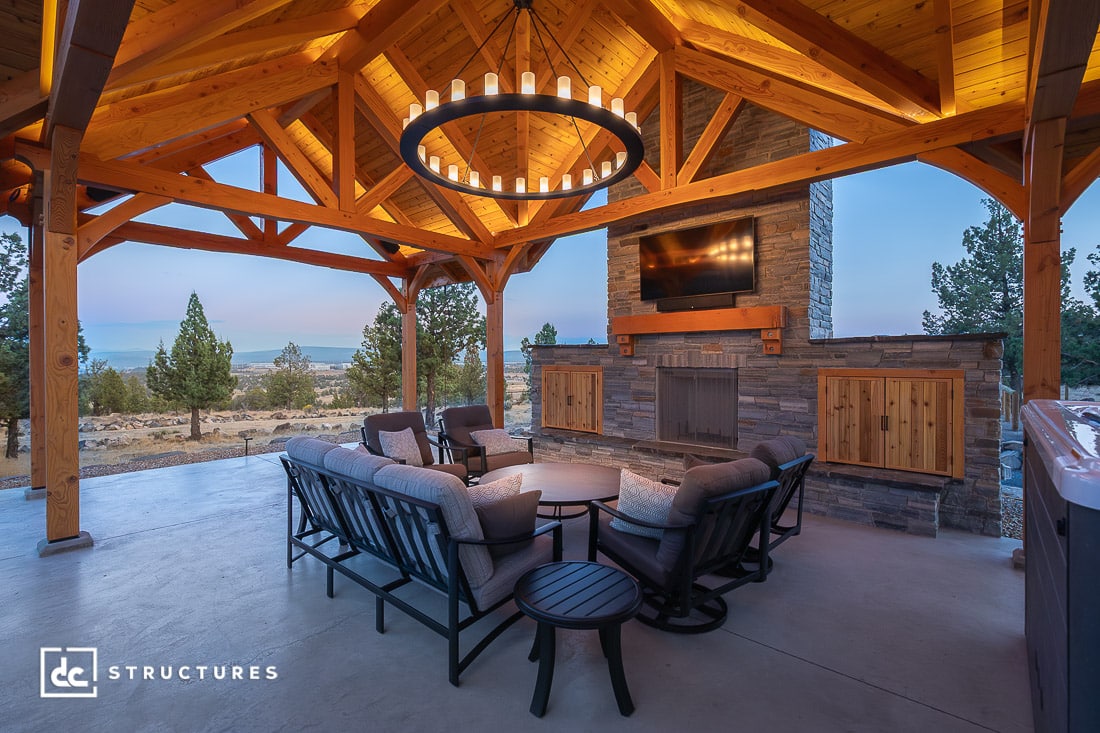 A covered outdoor patio with wooden beams, a circular chandelier, cushioned seating around a fire pit, and a stone fireplace at dusk.