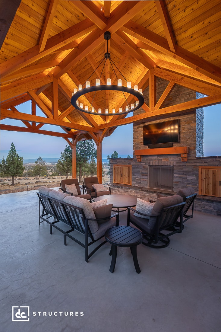 A cozy covered patio with cushioned seating around a fireplace and TV, wooden vaulted ceiling, and scenic windows showing hills.