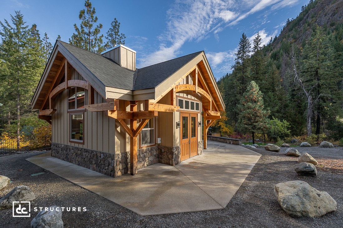 A modern cabin-style building with wood and stone siding, large windows, and a peaked roof sits among trees and mountains. Large rocks and a concrete path are in the foreground.