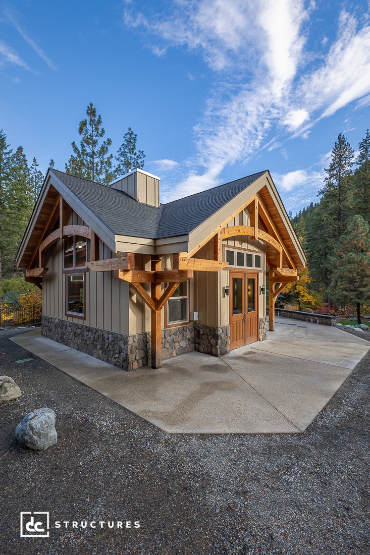 A modern cabin with wood beams, stone accents, and large windows sits on a concrete patio surrounded by trees and mountains under a partly cloudy sky.