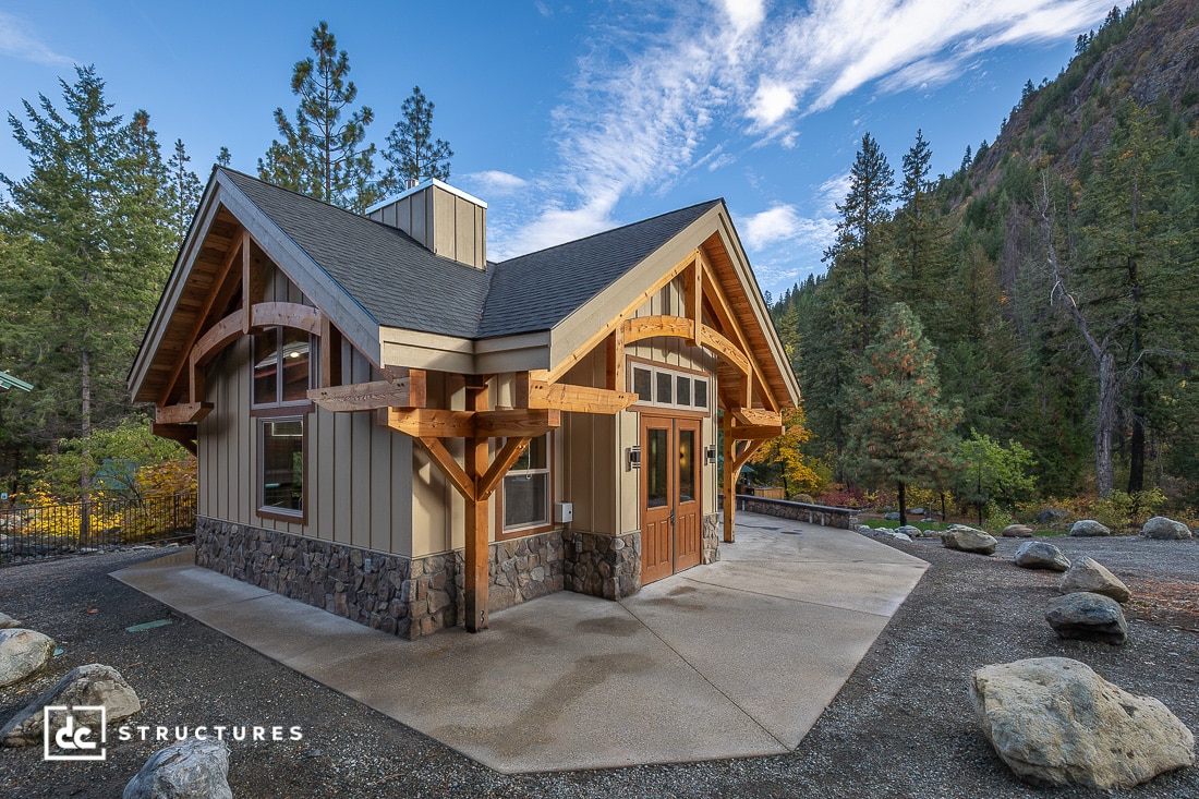 A small modern cabin with stone and wood siding, large windows, and a peaked roof sits among pine trees and mountains under a cloudy sky.