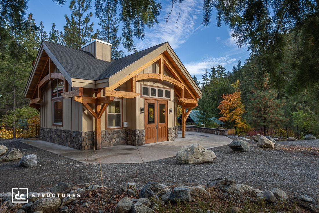 A small modern cabin with wooden beams and stone accents stands in a forested area with autumn trees and a clear sky overhead.