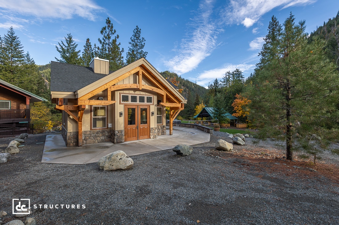 A small wooden cabin with stone accents sits among trees and mountains under a blue sky. Gravel, rocks, and a walkway surround it.