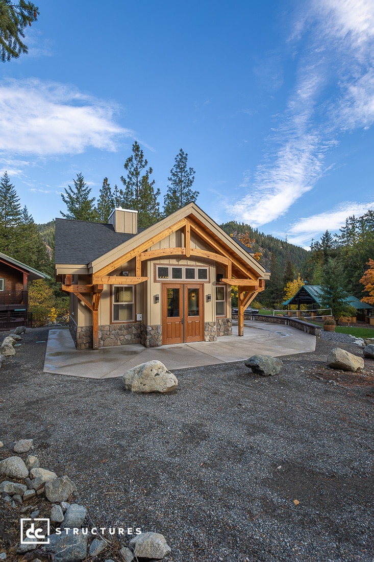 A small modern cabin with stone and wood exterior sits on a gravel lot among trees and mountains under a partly cloudy sky. A concrete path leads to the front double doors.