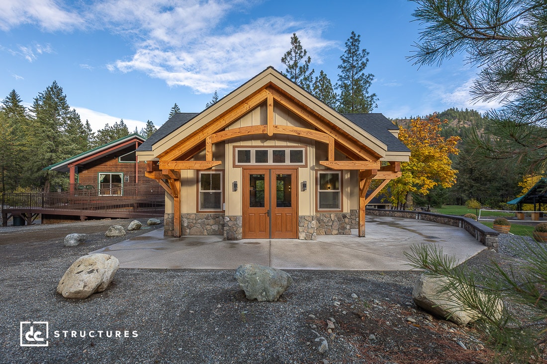 A small modern cabin with stone and wood accents stands in a wooded area, framed by large wooden beams and multiple windows.