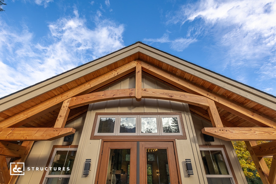 Close-up of a modern wooden house exterior with large windows and exposed timber beams, viewed from below against a blue sky with scattered clouds.
