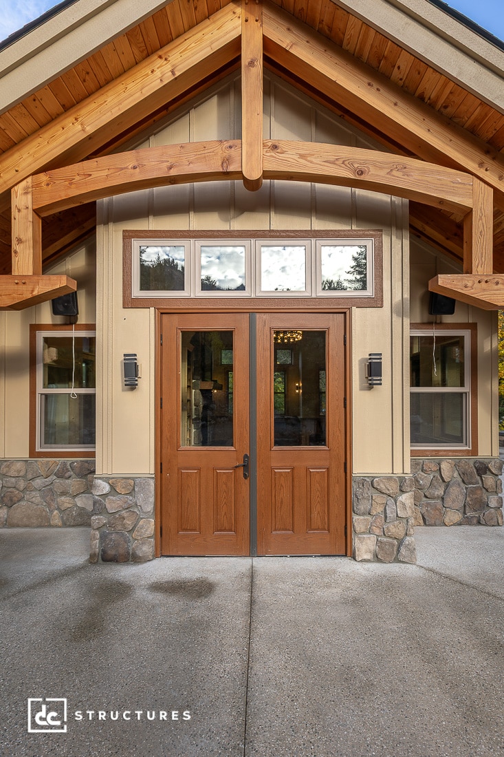 The image shows a house entrance with double wooden doors, stone accents, beige siding, a peaked porch roof, and four windows above.