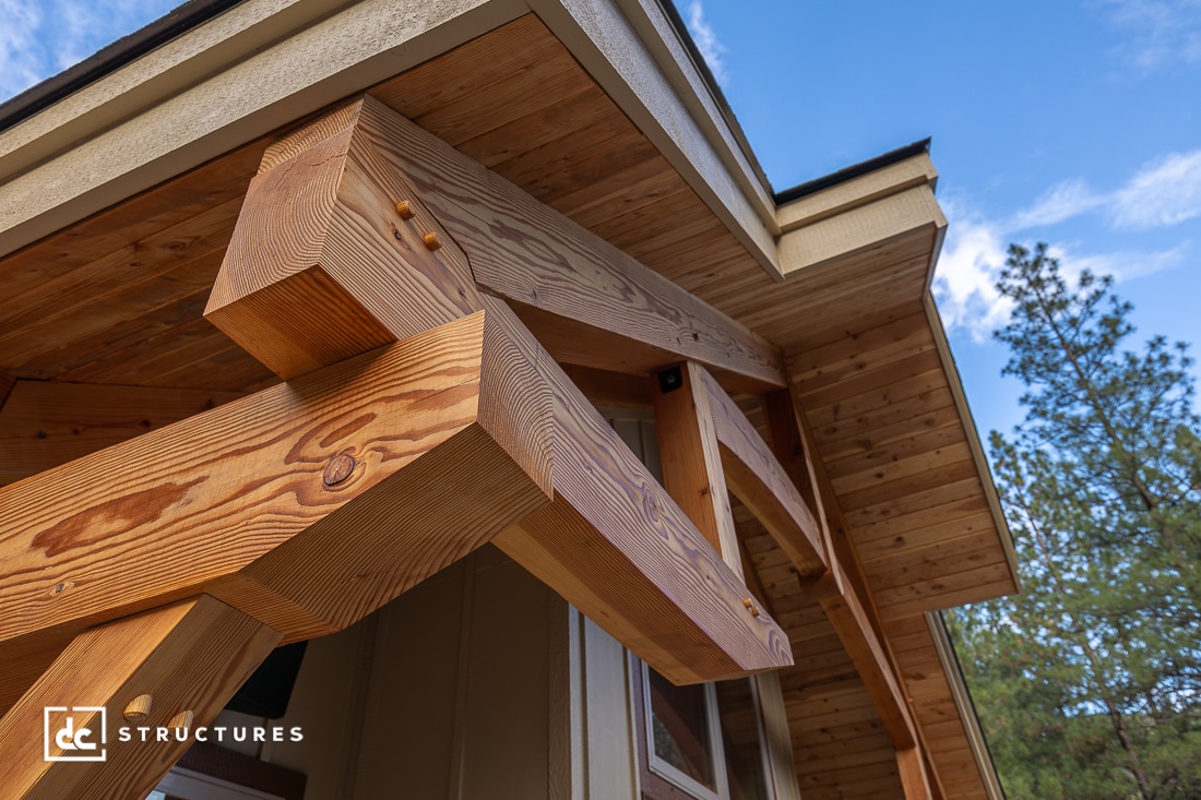 Close-up of a timber frame roof overhang with exposed wooden beams and detailed joinery, set against a blue sky with trees in the background.