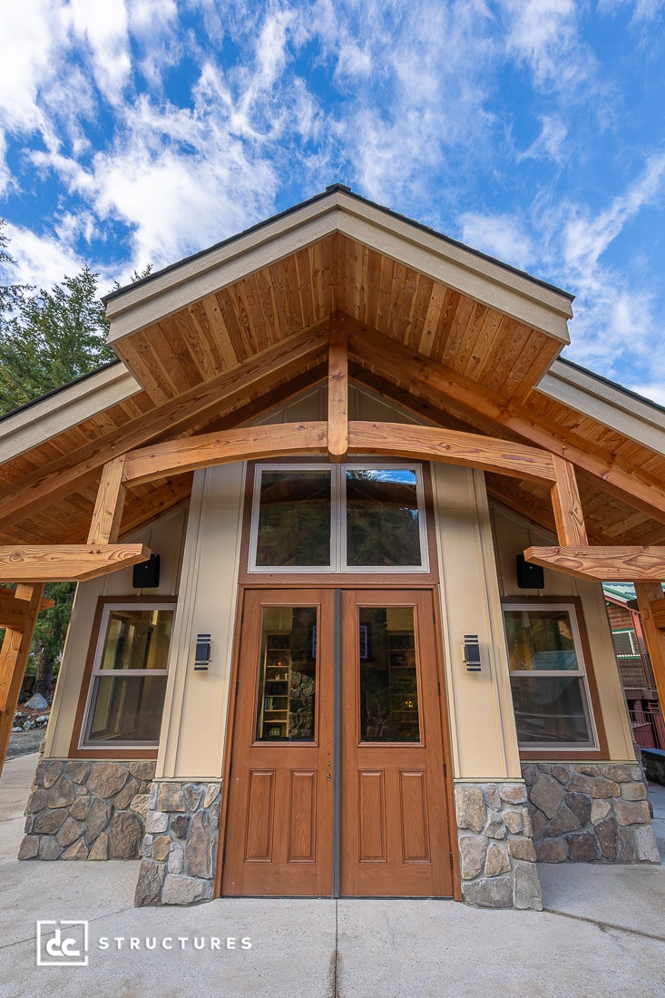 A modern building with a peaked wooden roof, large double wooden doors, stone accents at the base, and tall windows against a blue sky.