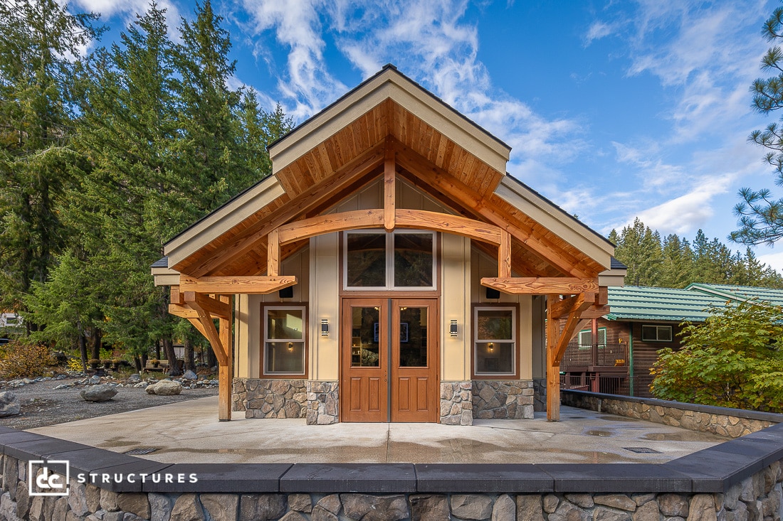 A small, modern cabin with wood and stone exterior, large windows, and a peaked roof, surrounded by trees under a blue sky.