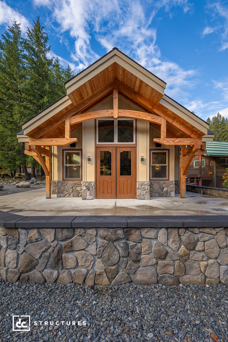 A modern cabin with a wooden frame, stone accents, large windows, and double doors on a stone terrace with trees and blue sky.