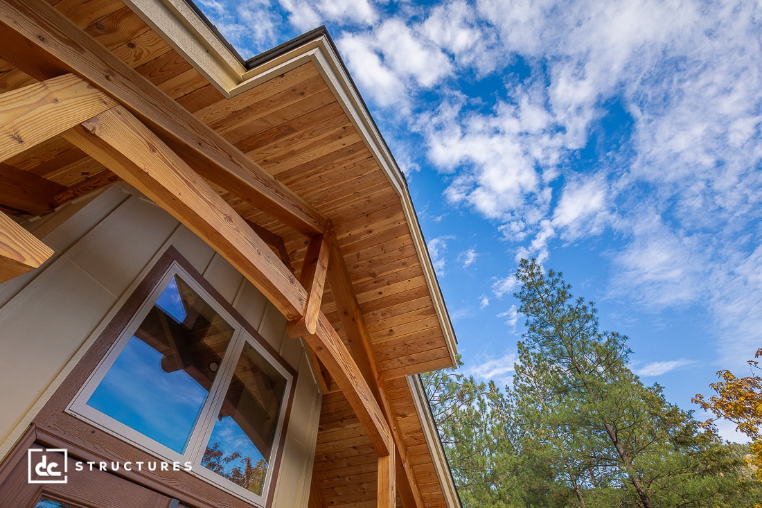 A modern wooden house exterior with large eaves and exposed beams, seen from below against a blue sky with scattered clouds and nearby pine trees.