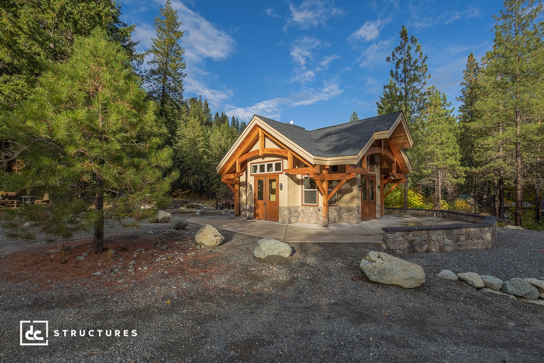 A small modern cabin with timber accents stands among tall pines and rocks under a blue sky, with forested hills in the background.