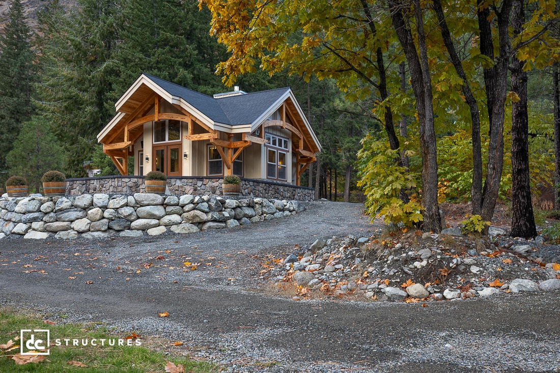 A modern wood and stone cabin sits on a raised stone foundation, surrounded by trees, gravel driveway, and autumn leaves.