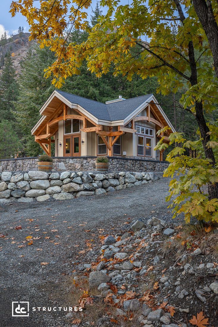 A small modern wooden cabin with large windows sits on a stone wall, surrounded by autumn trees and mountains. Gravel path leads up.