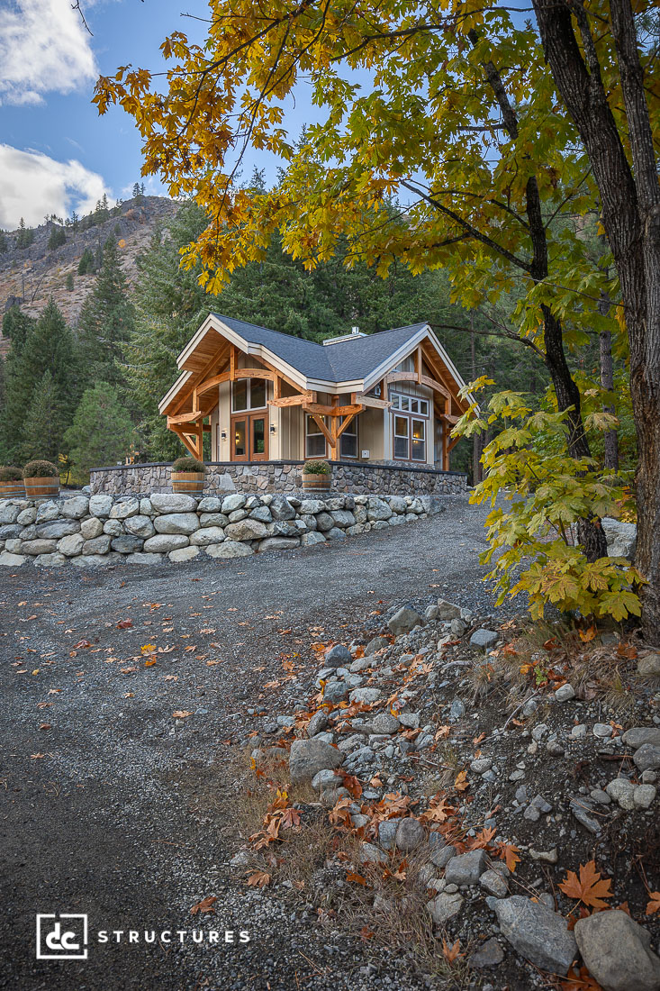 A modern wooden cabin with large windows sits on a stone foundation, surrounded by autumn trees and mountains. Fallen leaves cover the gravel driveway.