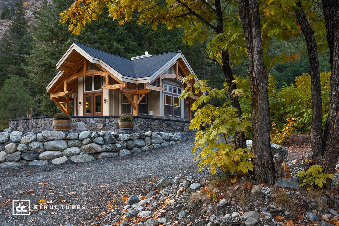 A small modern cottage with large windows and a stone foundation sits on a gravel driveway, surrounded by autumn trees and a forested hillside.